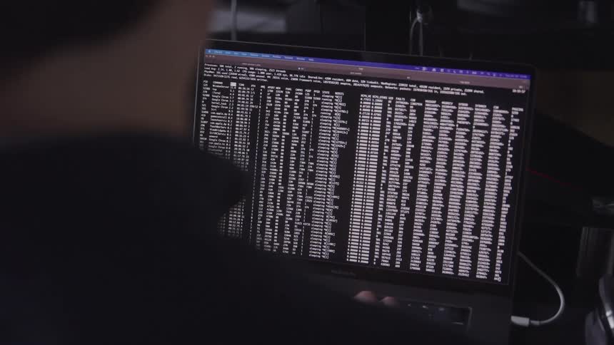 The man is intently viewing a laptop screen filled with extensive lines of code in a dimly lit environment meticulously focusing on data or programming details at his workstation