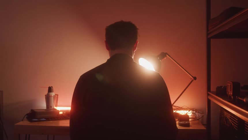 A man stands at a wooden workbench illuminated by a desk lamp with shelves of electronics visible on his right in a dimly lit setting