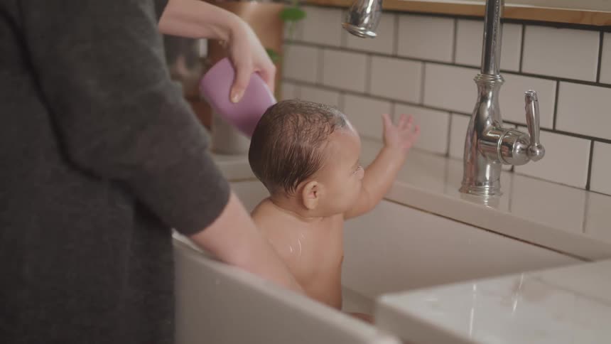 An adult is bathing a baby in a kitchen sink with the baby reaching towards the faucet