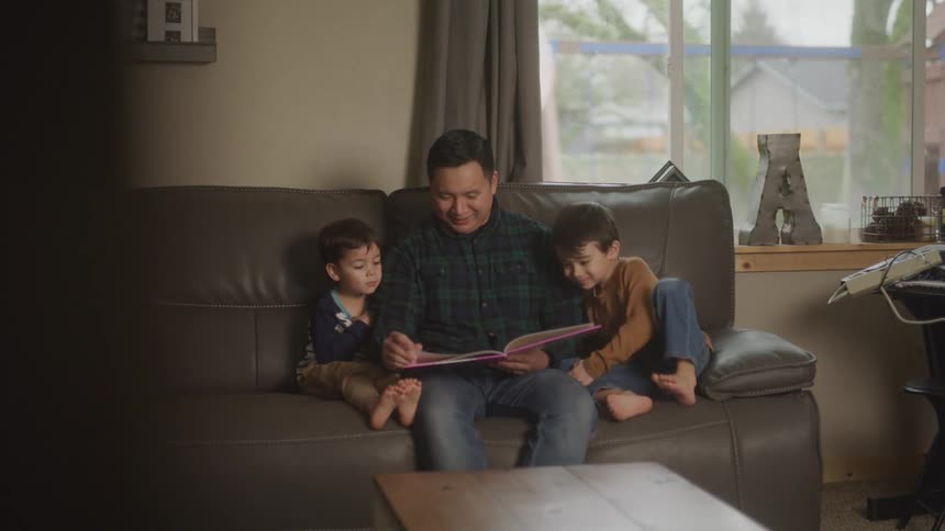 A person is sitting on a couch reading a book to two children in a cozy living room with a window showing a rainy day outside
