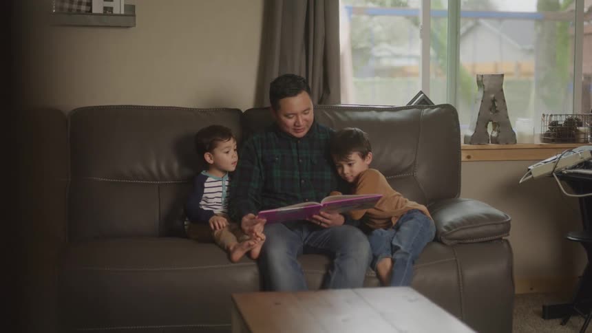 A father sits comfortably on a brown leather couch in a living room reading a book aloud to his two young sons who are nestled beside him