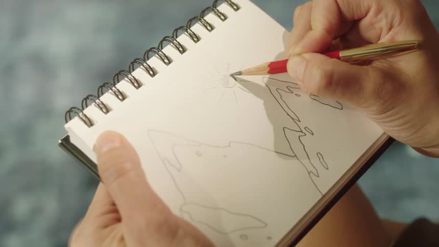 A person is holding a spiral bound sketchbook and drawing a simple outline of a figure with a red and gold colored pencil