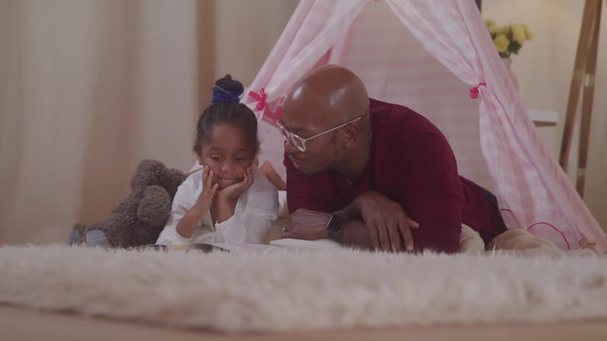 Father with a shaved head and glasses is lying on a fluffy white rug inside a pink play tent looking at a book with daughter who has dark hair styled in a bun with a blue scrunchie and is resting her chin on her hands