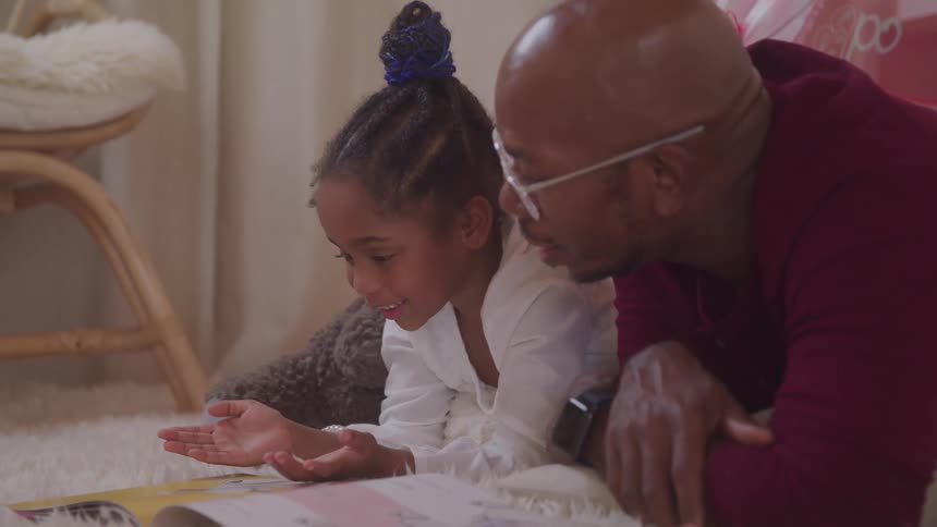 Laying sidebyside on a soft rug indoors a young girl gestures with her hands while talking to her father as they look together at an open book spread before them
