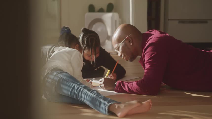 A father draws with his daughters while they lie on the living room floor