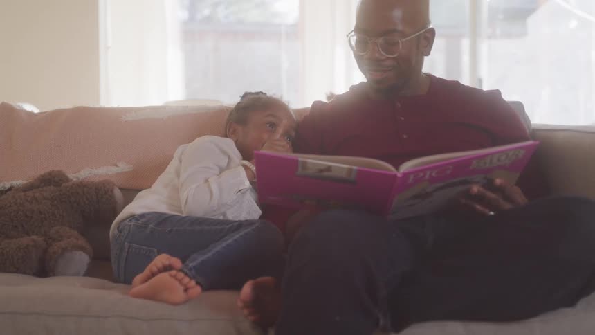 A father reads a book to his daughter while they sit on the living room sofa