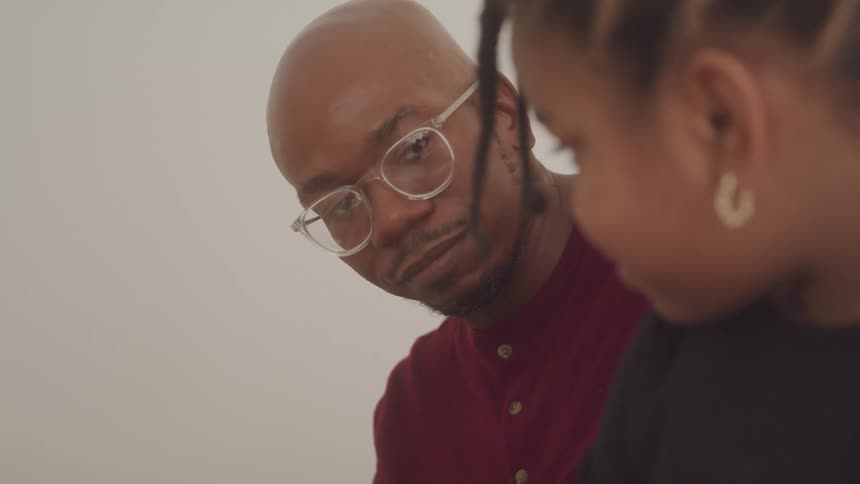 A closeup shot captures a tender moment indoors as a young girl with braided hair turns her head to look directly at her attentive father who is wearing glasses