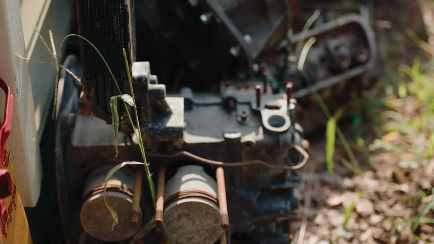 A closeup shot reveals weathered pistons and parts of a discarded motor lying abandoned on the dirt ground outdoors with blades of grass nearby
