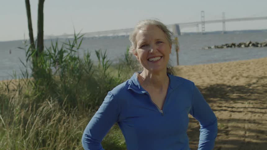 An older woman smiles while standing on a sandy beach on a sunny summer day