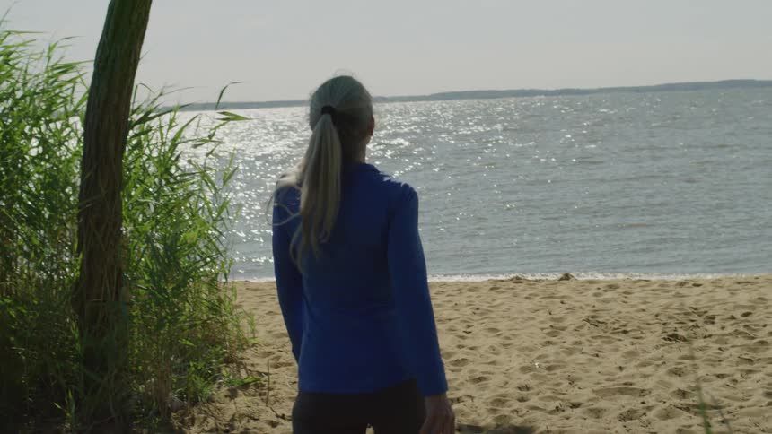 Wearing a blue longsleeve shirt and dark leggings a woman walks into frame towards the sandy shoreline where sunlight sparkles brightly on the gently waving water