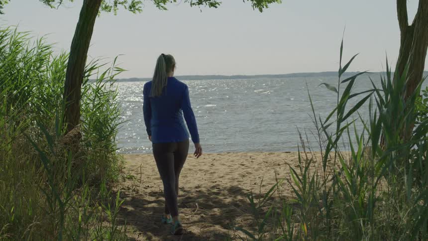 A woman in athletic wear walks out from between trees and grass onto a sandy shore stopping to gaze at the sunlit water