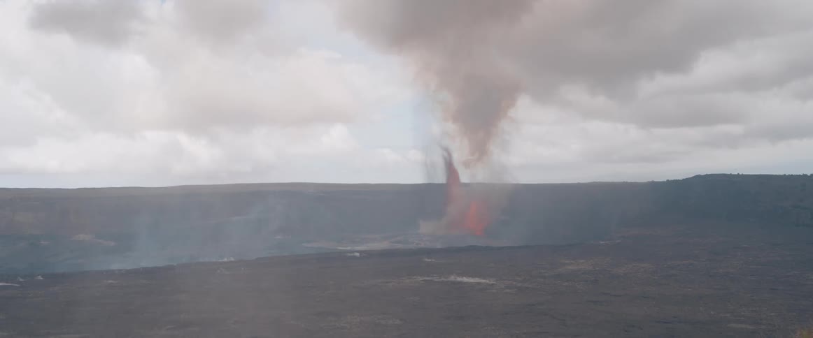 A wide shot shows a massive volcanic crater with dark solidified lava fields as a plume of smoke and ash rises high from an active lava fountain erupting in the distance