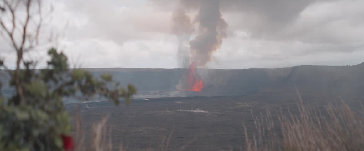 Viewed from an overlook with sparse vegetation in the foreground a powerful lava fountain erupts within a vast volcanic crater sending a thick plume of smoke and ash into the cloudy sky