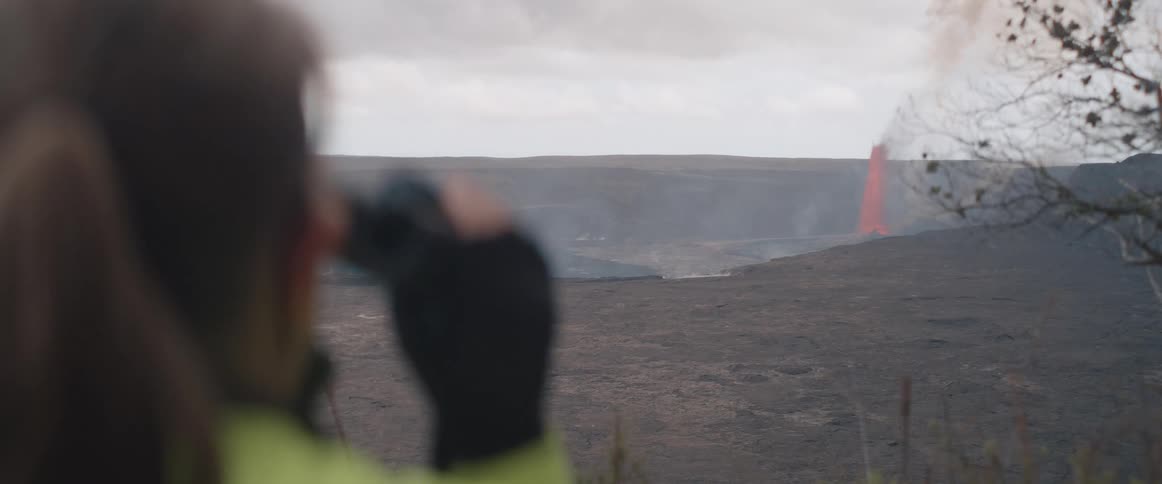 Seen from over the shoulder a person wearing gloves raises binoculars to their eyes to watch an active volcanic eruption with a visible lava fountain in the distant crater