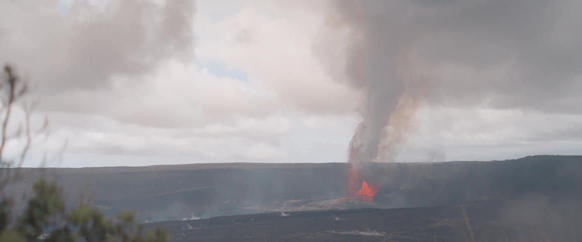 A volcano erupts forcefully sending lava fountaining high into the air and emitting a large plume of smoke against a cloudy sky over a dark barren landscape