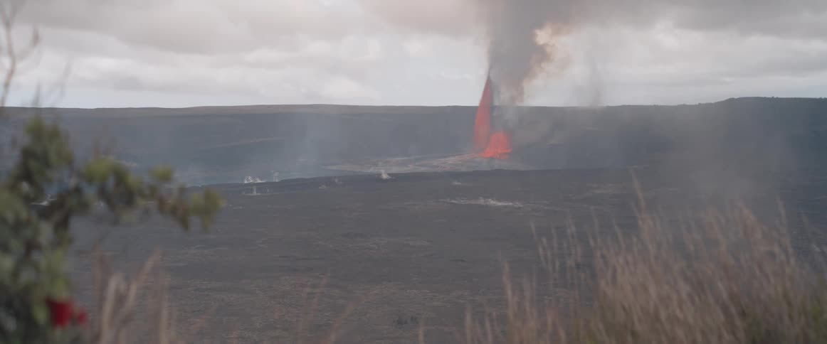 From a distance an active volcano erupts with a bright orange lava fountain spewing molten rock and a large plume of smoke rising from the vast crater floor under a cloudy sky