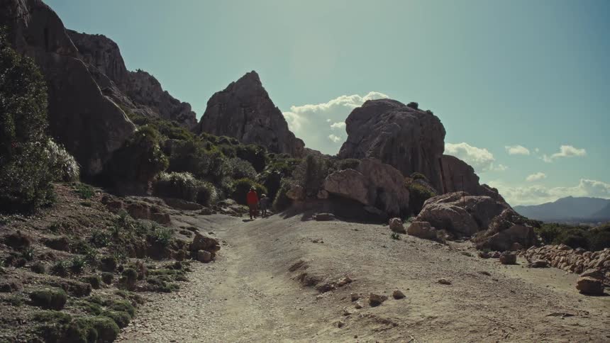 A woman and child hike away from the camera along a dirt trail through a sunny arid landscape dominated by large rock formations and sparse vegetation