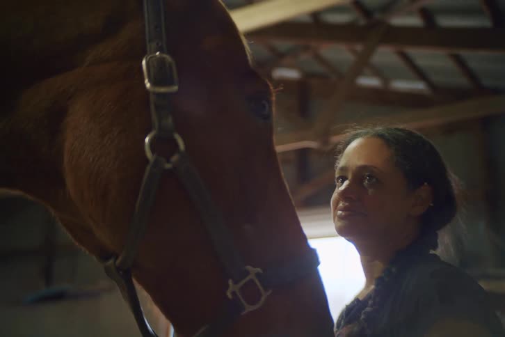 Inside a dimly lit stable a woman with braided hair smiles warmly while looking up at the head of a brown horse standing close beside her