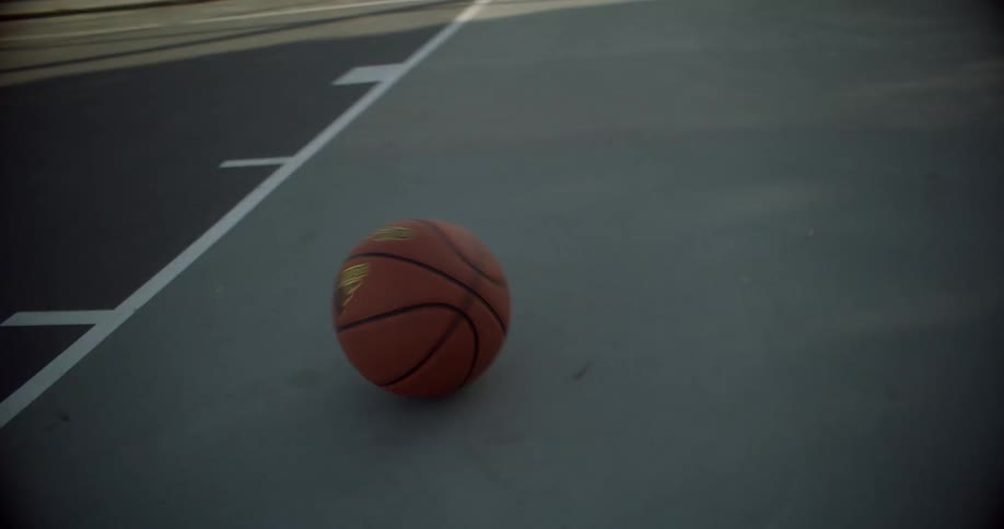 An orange basketball rolls across the grey surface of an outdoor court moving towards and alongside painted white boundary lines