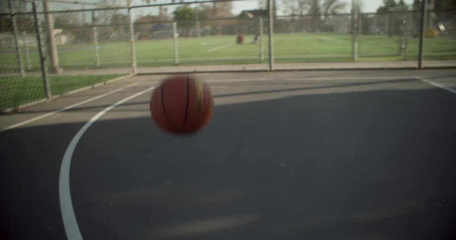Captured with motion blur an orange basketball bounces rhythmically on the grey surface of an outdoor court with the hoop and chainlink fence visible beyond