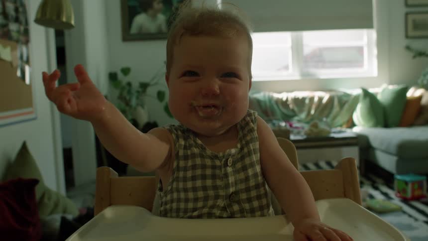 A baby wearing a checkered outfit sits in a high chair with their arms raised in a brightly lit room