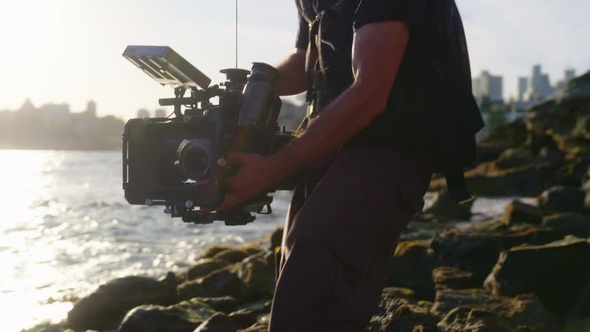 A filmmaker holds his camera while filming on a rocky lakeshore near a metropolis