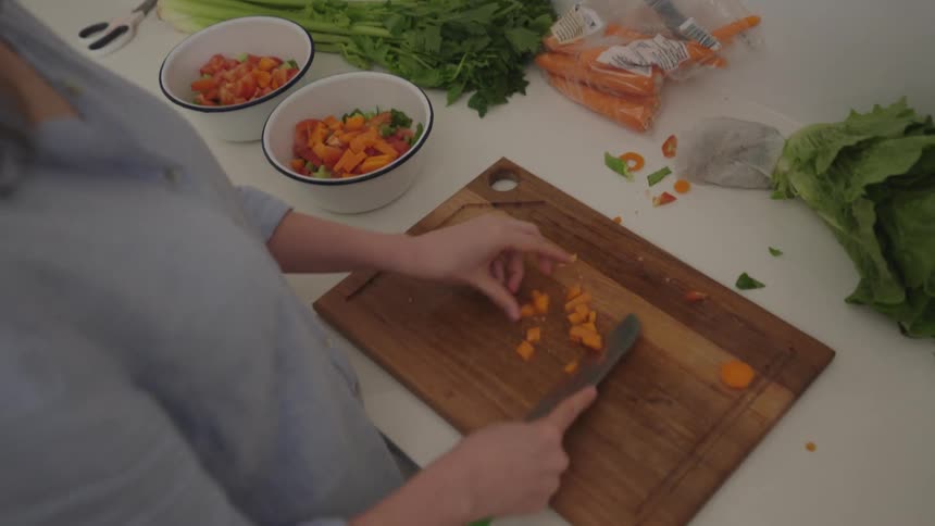 A woman in a light blue shirt is preparing food on a white countertop transferring chopped orange bell peppers from a wooden cutting board into a white bowl containing diced tomatoes