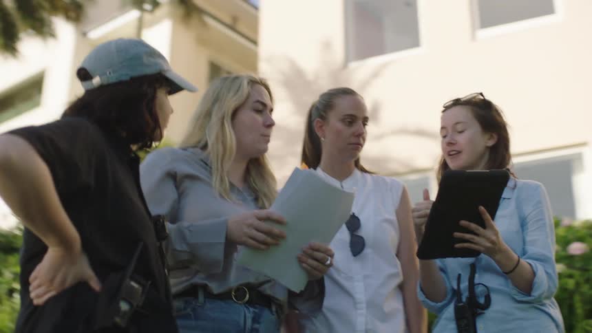 Four young women are standing outdoors next to an apartment building while looking at an electronic tablet