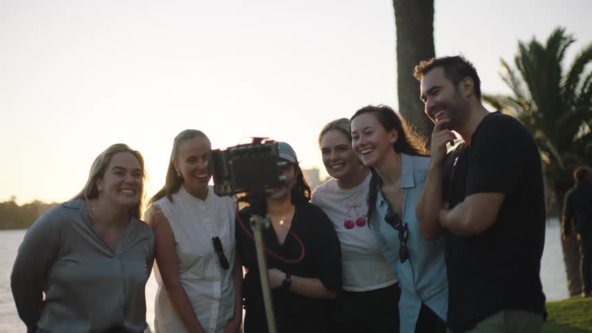 Six people mostly women facing a camera interacting happily