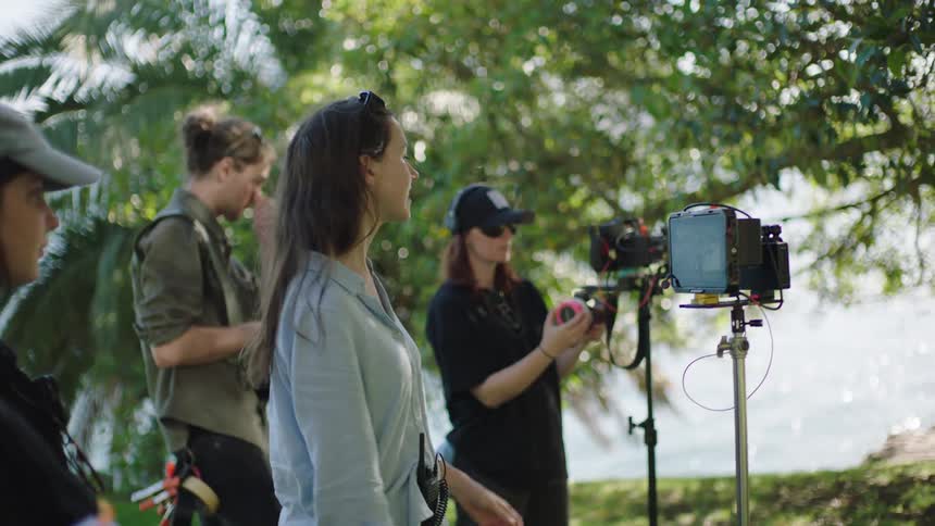 A film crew sets up equipment outdoors featuring a camera mounted on a tripod against a backdrop of lush greenery