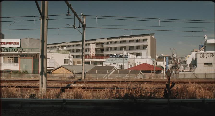 Railway tracks beneath overhead power lines surrounded by buildings with japanese signs indicating a hotel and school in the background
