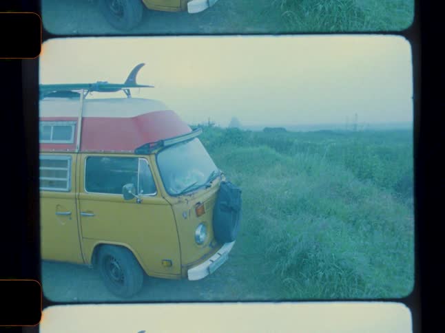 A yellow vw camper van with a surfboard is parked in a hazy grassy landscape