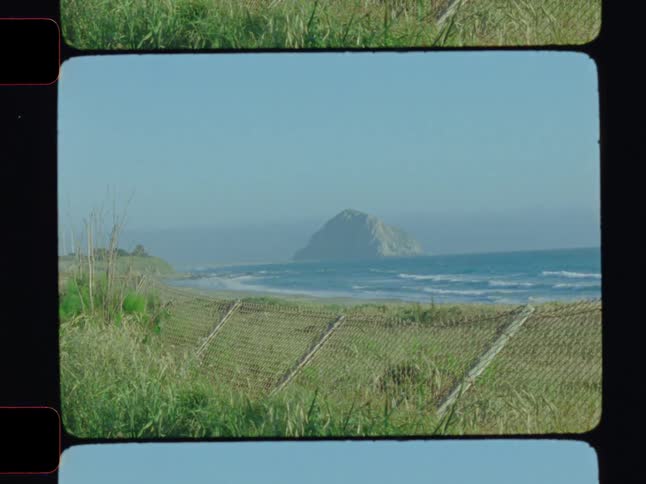 A tranquil coastal scene where a bended wire fence runs through tall grass in the foreground beyond which lies a sandy beach meeting gentle waves and a hazy headland under a clear light blue sky