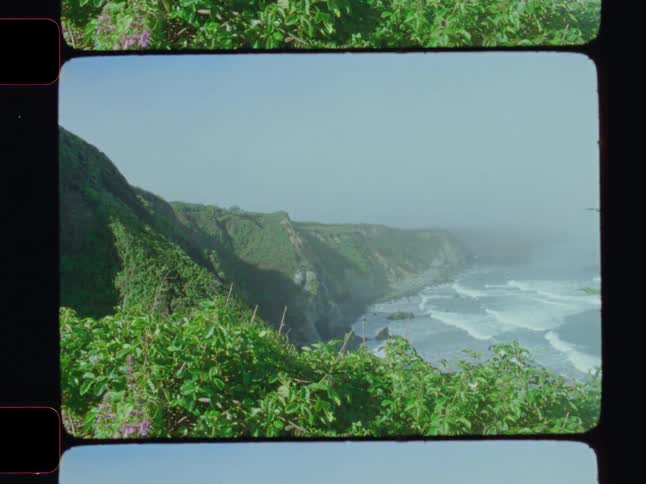 A scenic vista of a rugged green coastline with steep cliffs dropping down to crashing waves under a hazy light blue sky