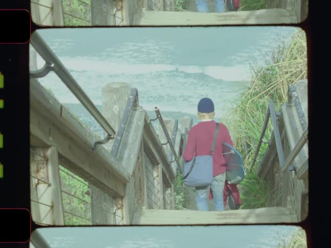 A person carrying a bag and surf board walks down wooden stairs towards the ocean waves