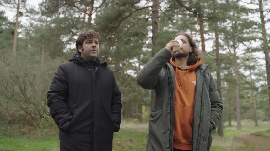 Two smiling men talking outdoors with one of them drinking from a disposable cup while standing in a forest