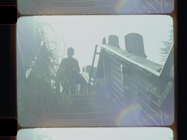 Viewed from below within a film frame a silhouetted person walks down outdoor wooden stairs against a bright hazy sky