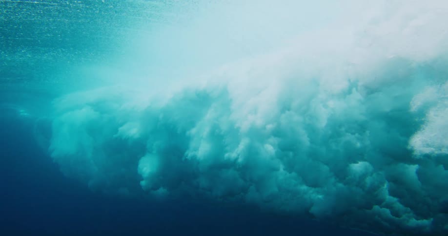 An underwater perspective of the powerful surge of a breaking wave filled with churning white foam and bubbles against the clear light blue water near the surface and the darker depths below