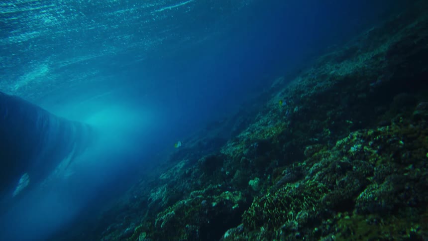 Underwater footage pans across a dark textured seabed covered in coral formations with sunlight filtering down through the deep blue water