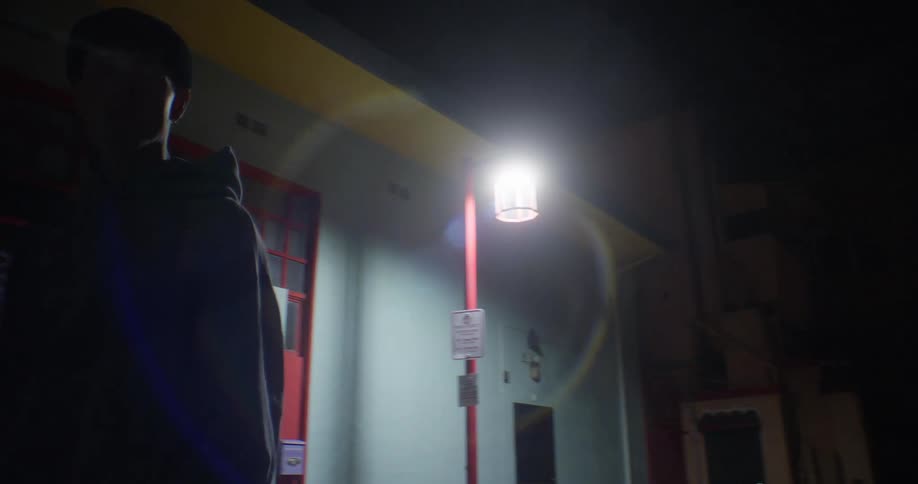 A person wearing a hoodie walks back and forth under the bright glare of a streetlamp in a dark urban alleyway at night