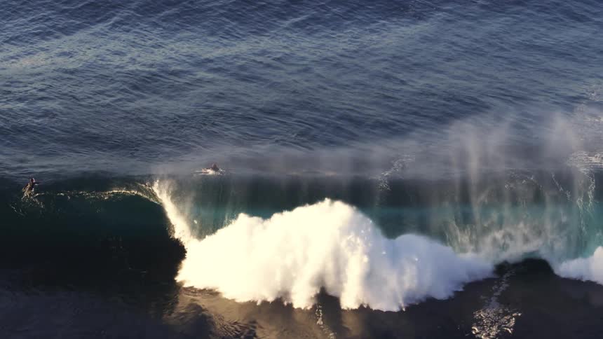 An aerial shot captures a surfer riding a large wave as it breaks dramatically across the dark ocean
