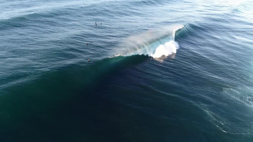 Surfers float in the water while a breaking wave creates a rainbow during the daytime