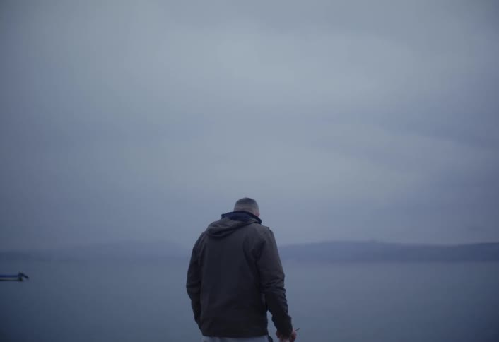 A man smoking a cigarette and holding a coffee cup turns his back to the camera to look out over the water under a grey overcast sky