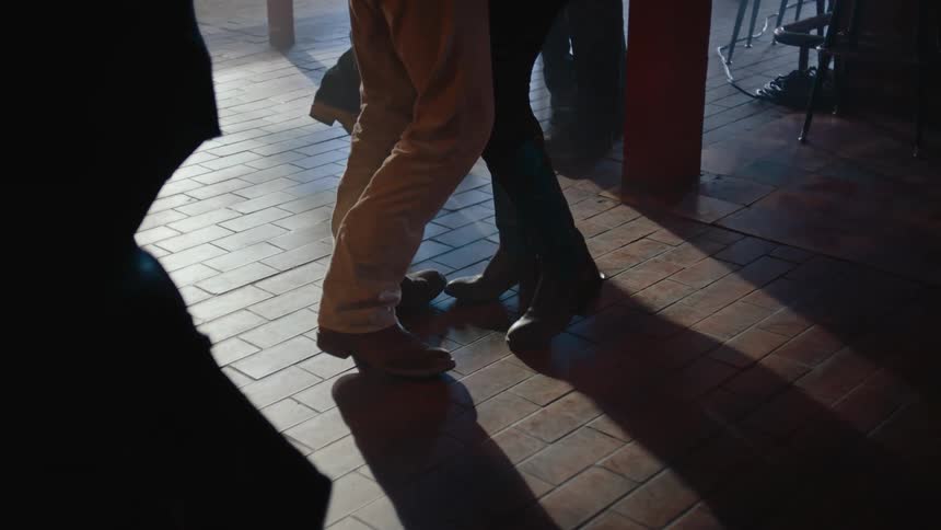Legs and feet move in a slow dance across a dimly lit brick floor inside a bar or club