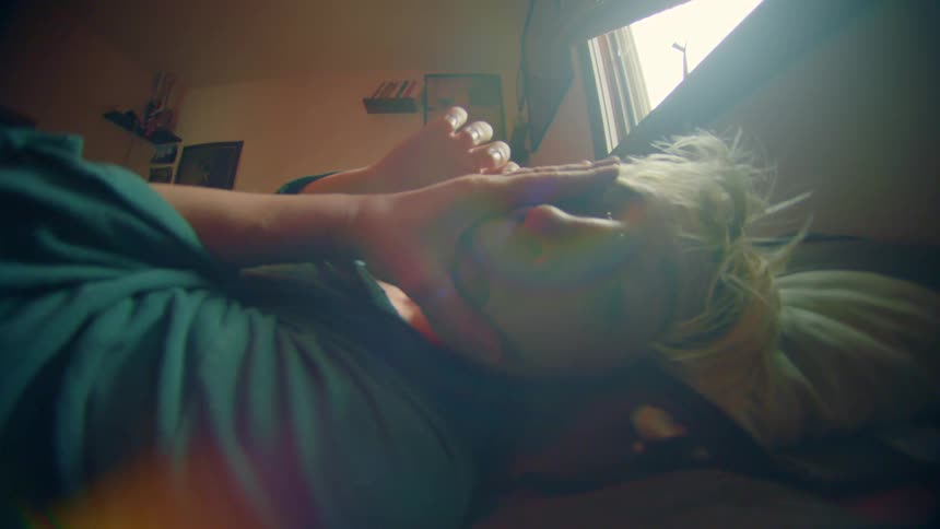 A sleepy blonde woman rolls in bed shielding herself from the morning light streaming through the window
