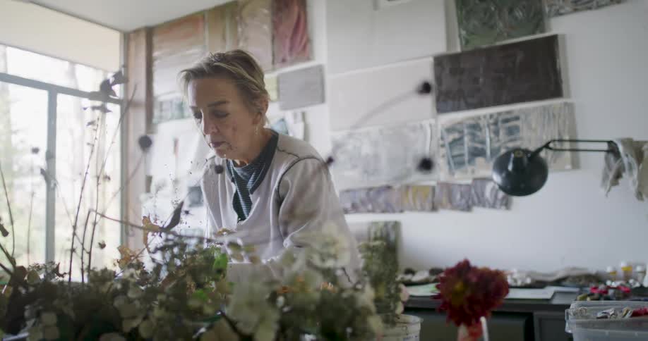 A female artist paints in her bright studio surrounded by artwork with dried flowers prominently arranged in the foreground