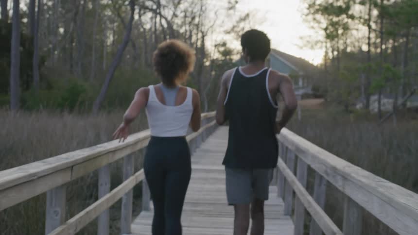 A man and woman are jogging across a wooden footbridge at sunset