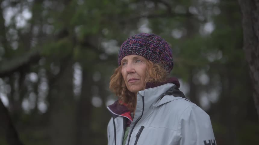 Woman wearing a knit hat and a jacket stands in the forest and looks around during the daytime