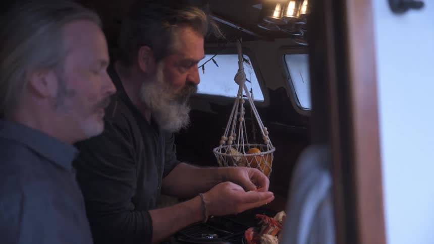 Two men enjoy eating crab in a small cramped kitchen aboard a boat sharing laughter and having fun