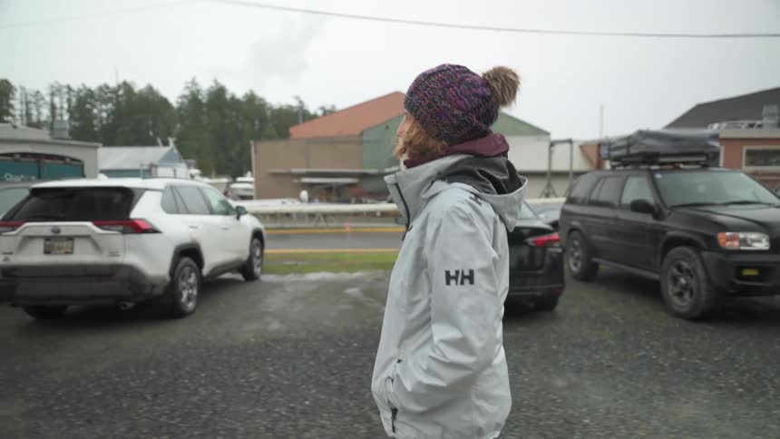 A woman wearing a winter hat and a grey jacket walks through a parking lot filled with cars on an overcast day
